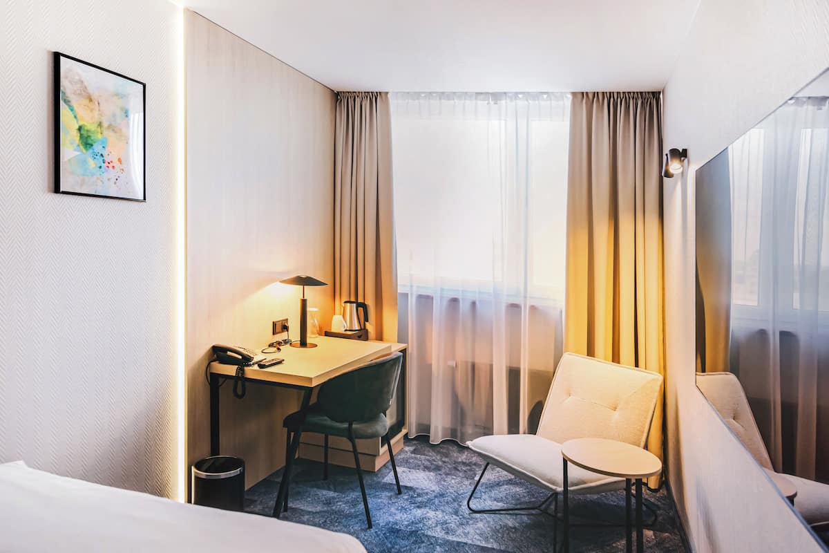 A hotel room with a bed, a desk with a phone and lamp, a chair, and a view through sheer curtains.