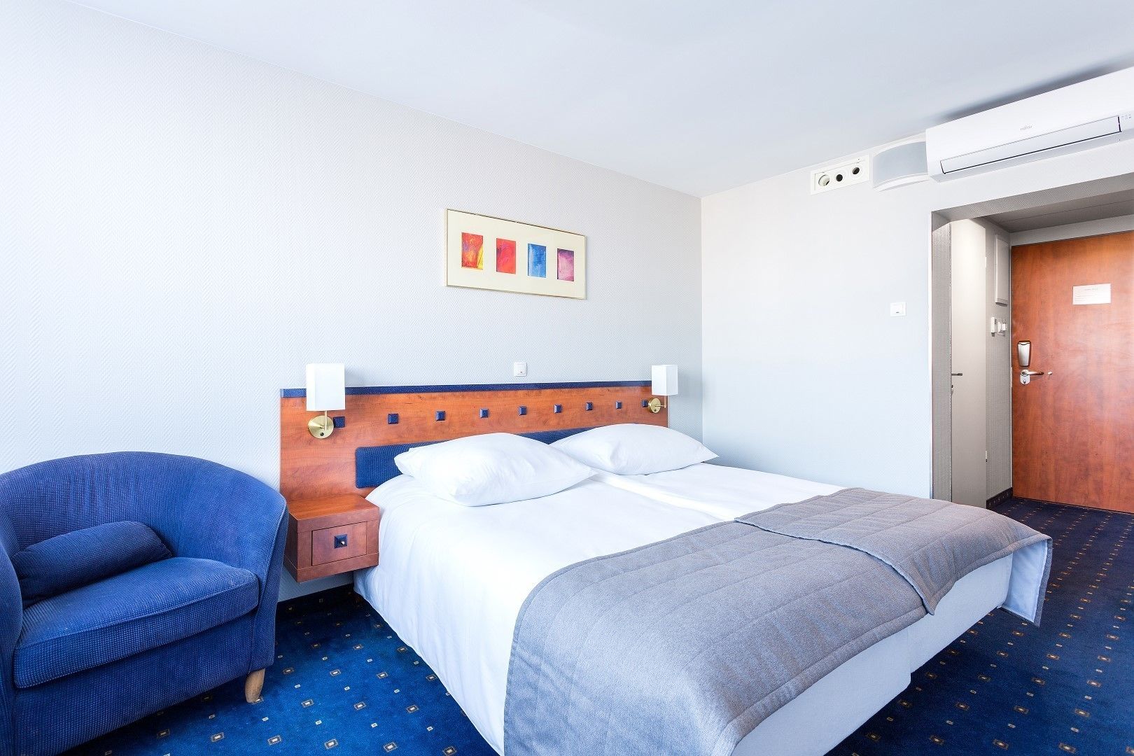 A hotel room with a bed, a chair, a nightstand, a wall-mounted air conditioner, and a door.