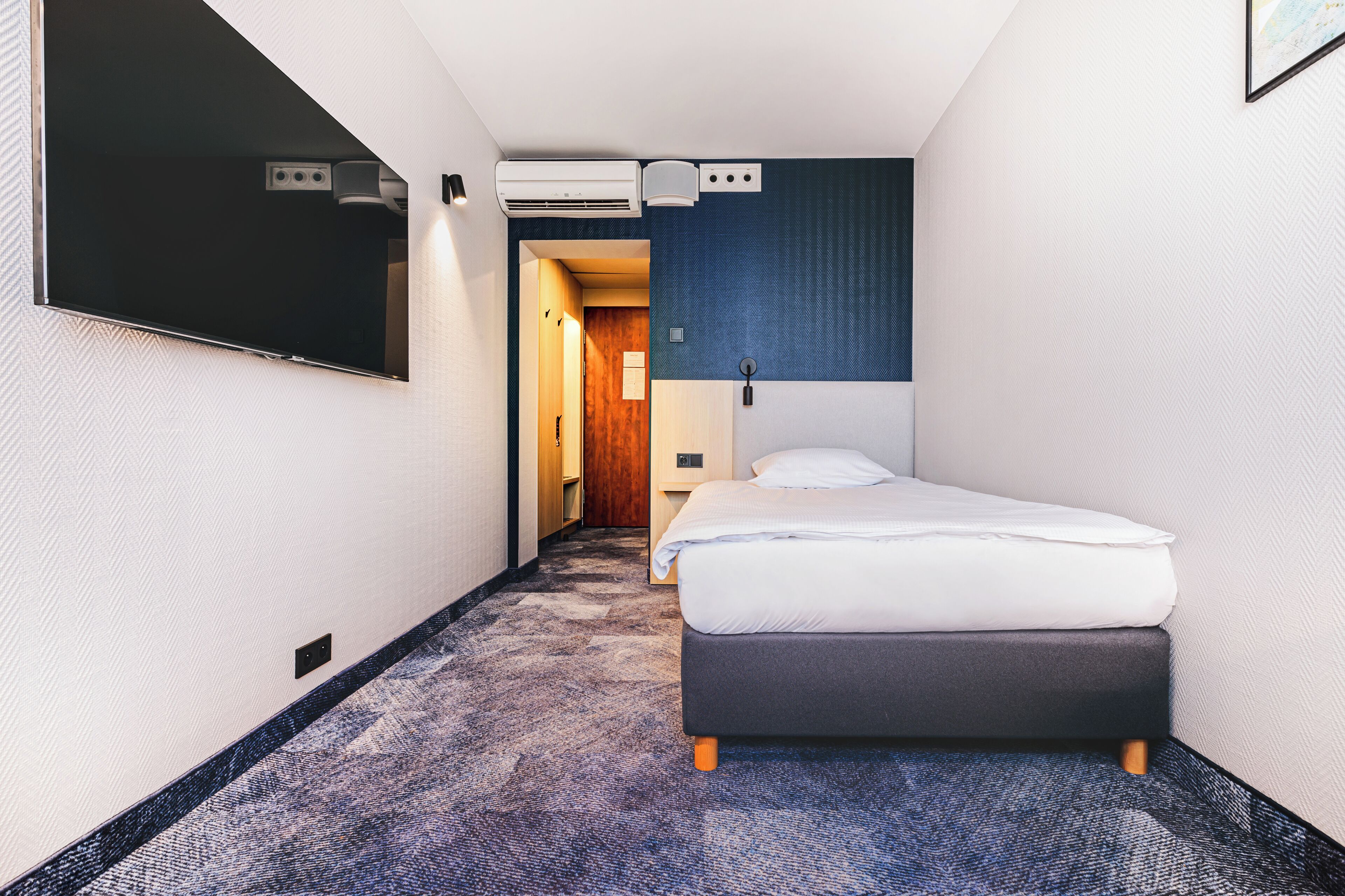 Camera d'albergo con un letto, una TV a schermo piatto montata a parete, un condizionatore d'aria e un pavimento rivestito di moquette.