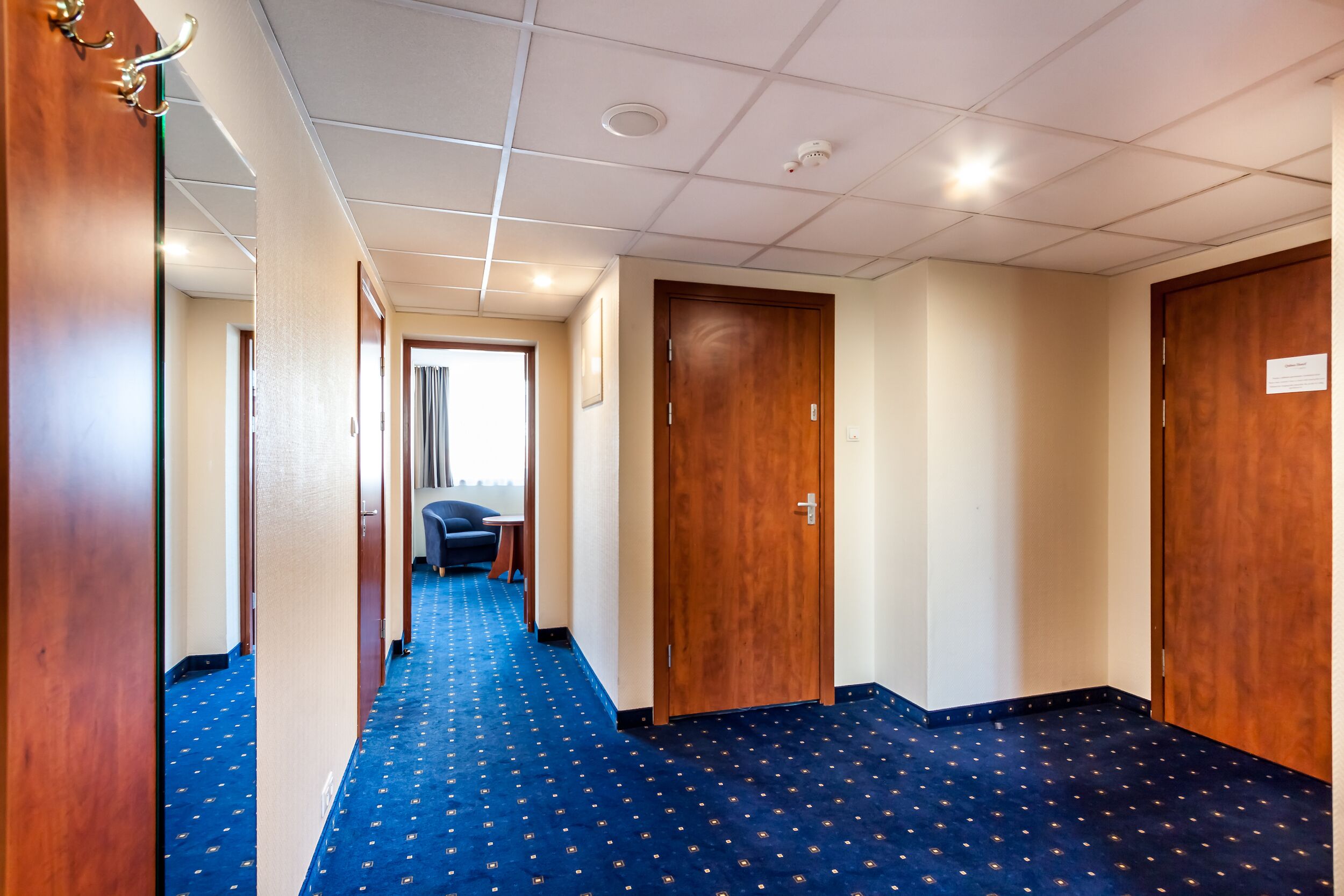 Pasillo de hotel con alfombra azul, puertas de madera y vista a una habitación con escritorio y silla.