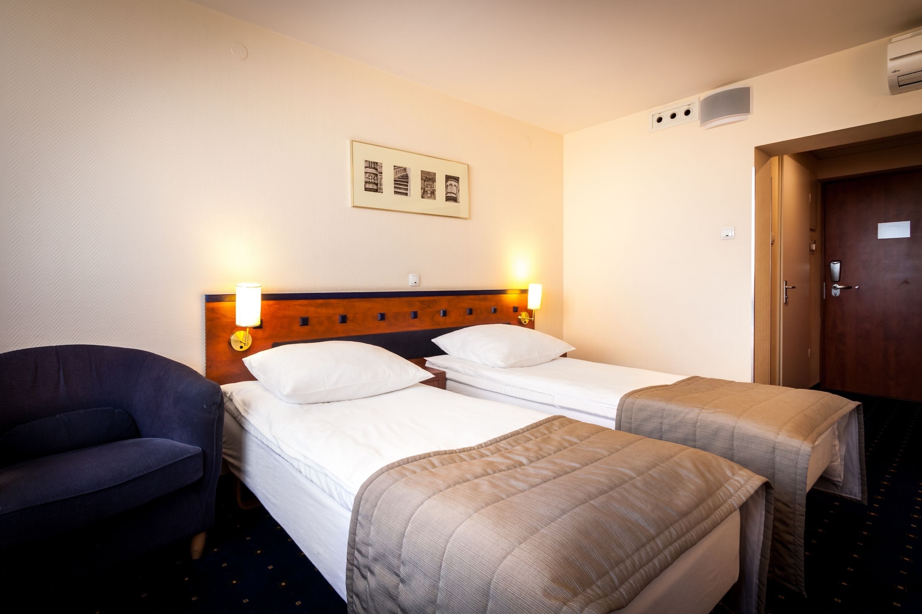 A hotel room with two single beds, a wooden headboard, a wall-mounted picture, a desk with a chair, and a door with a handle.