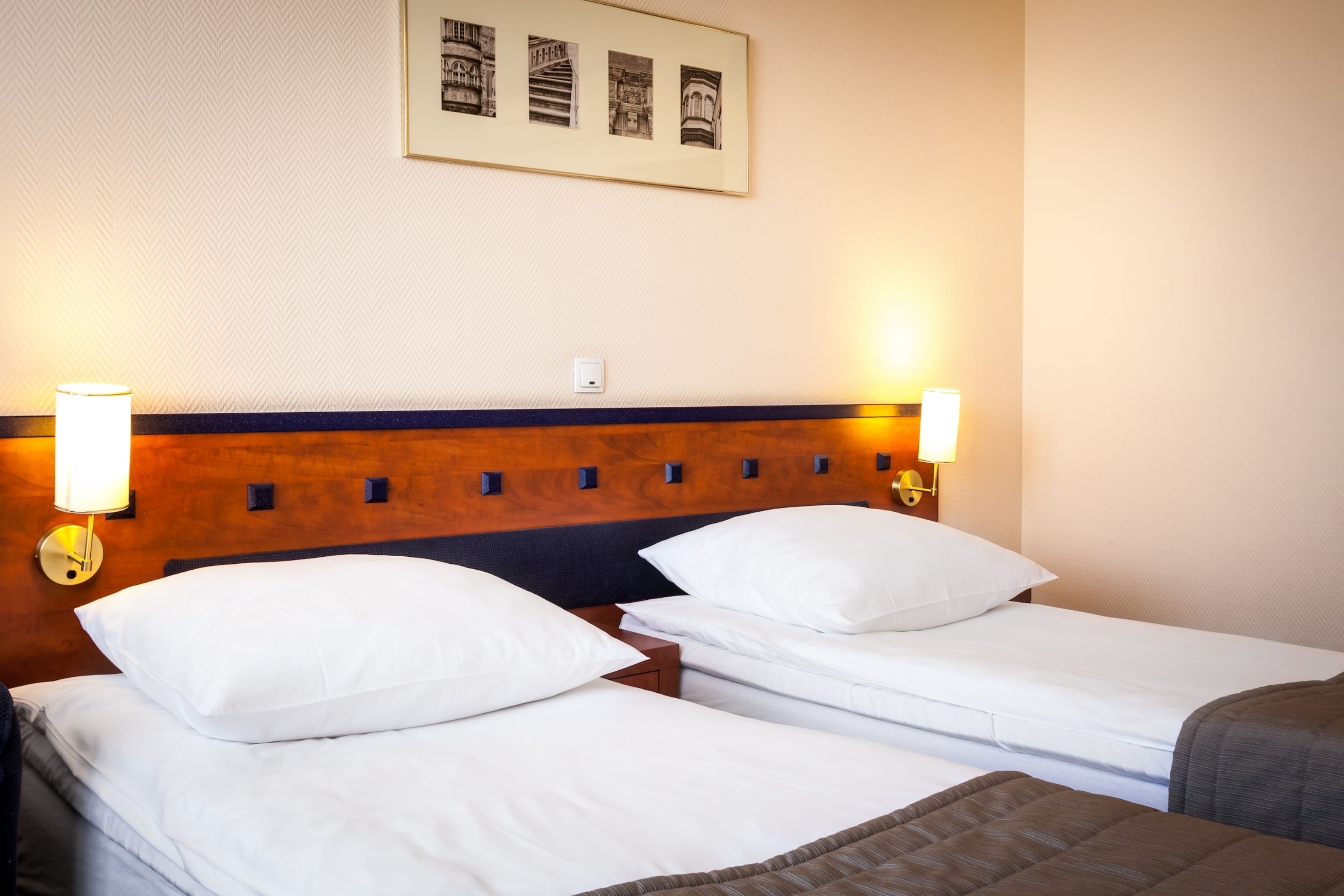 Ett hotellrum med två separata sängar, båda med vita sängkläder och kuddar, ett träfärgat sänggavel och väggmonterade läslampor.