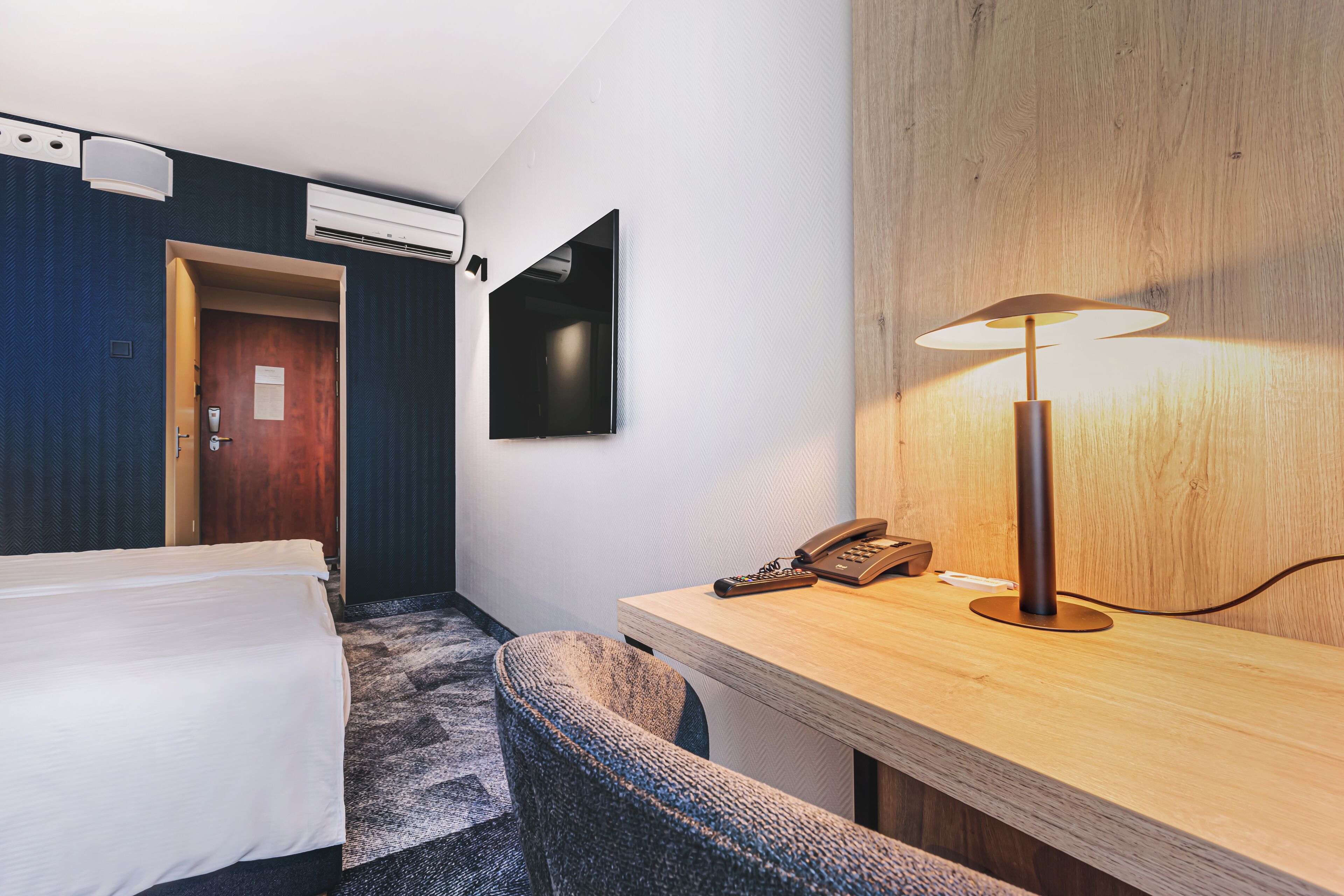 A hotel room with a bed, a desk with a lamp, a telephone, and a television on the wall.