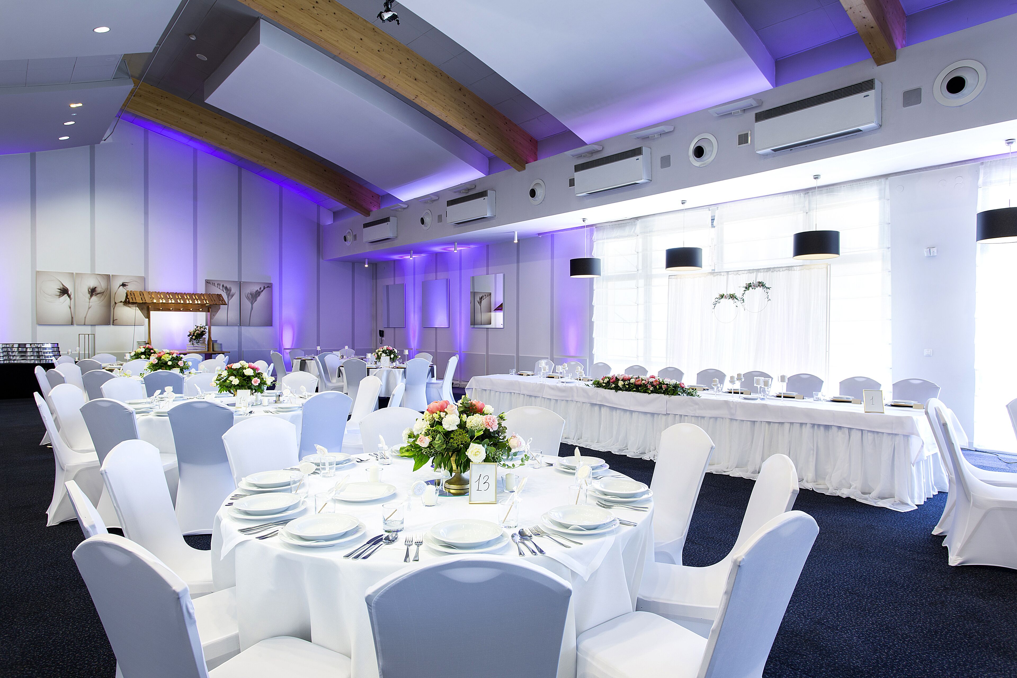 Une salle de banquet avec des tables rondes, des chaises blanches et des centres de table ornés de compositions florales.