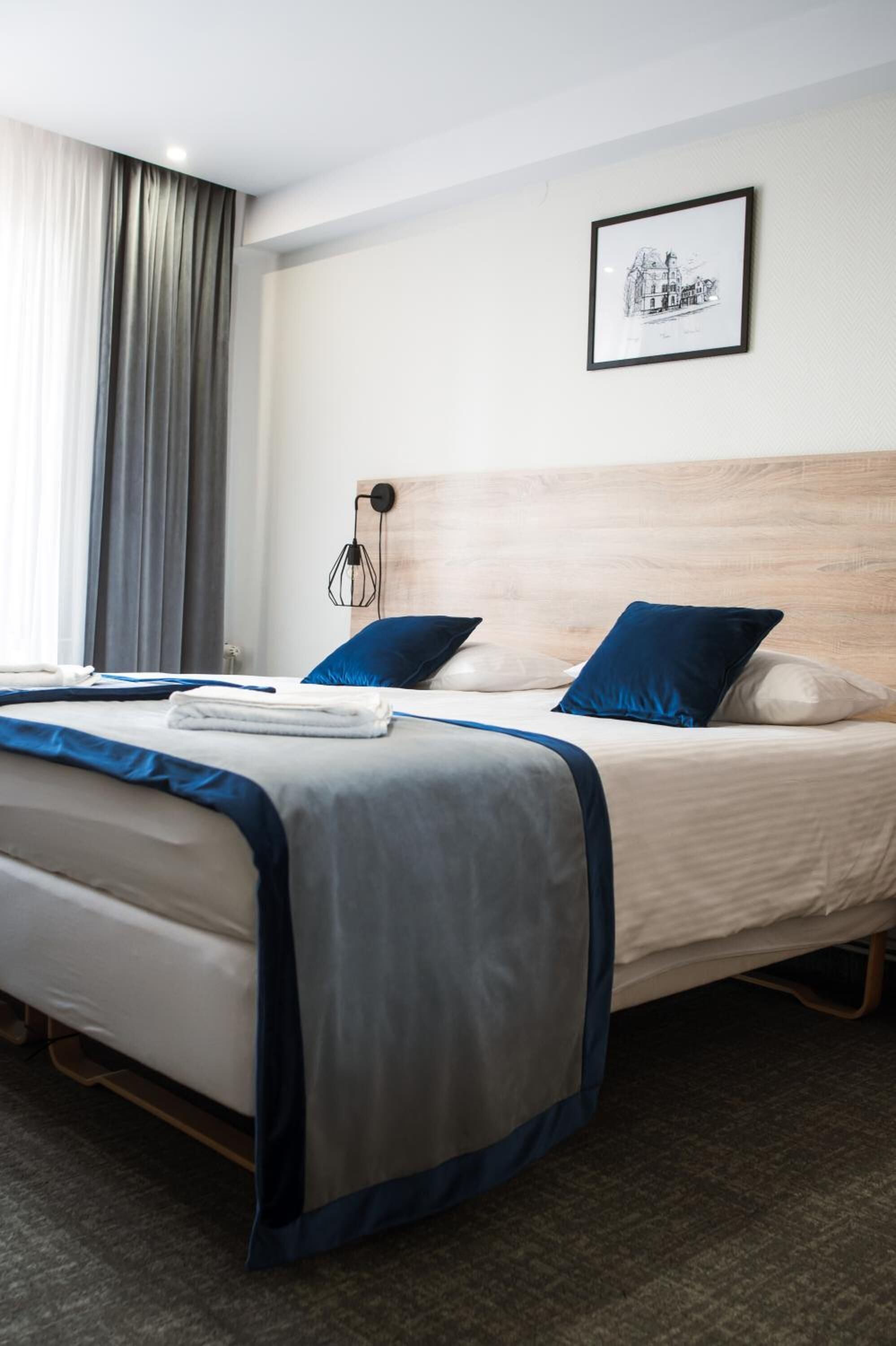 A hotel room with two beds, a large window with curtains, a framed picture on the wall, and a bedside table with a lamp.