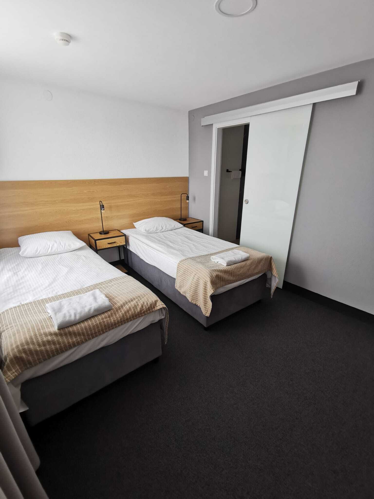 Habitación de hotel con dos camas, cabecero de madera, mesita de noche con lámpara y armario con puerta corredera.