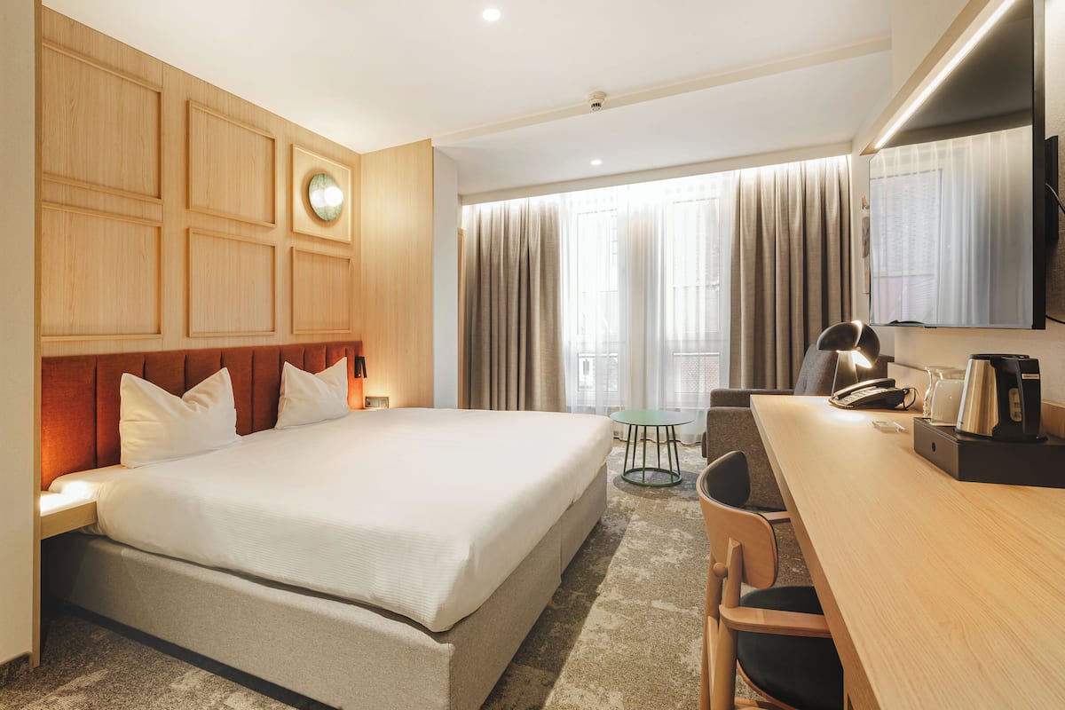 A hotel room with a large bed, a desk, a chair, a small table, a clock, and a television.