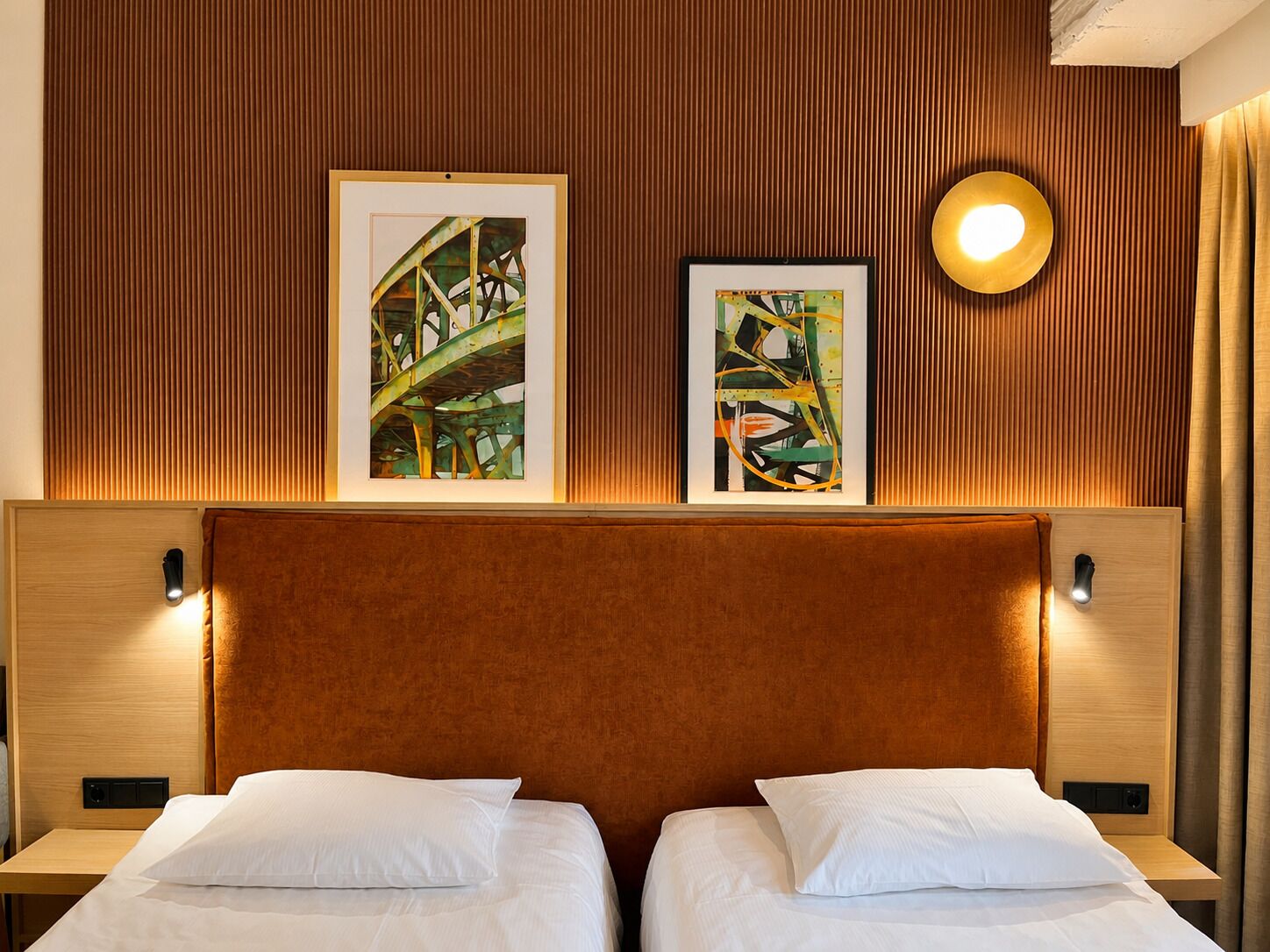 Une chambre d’hôtel avec une tête de lit en bois, deux lits avec du linge de lit blanc et deux œuvres d’art encadrées au-dessus des lits.