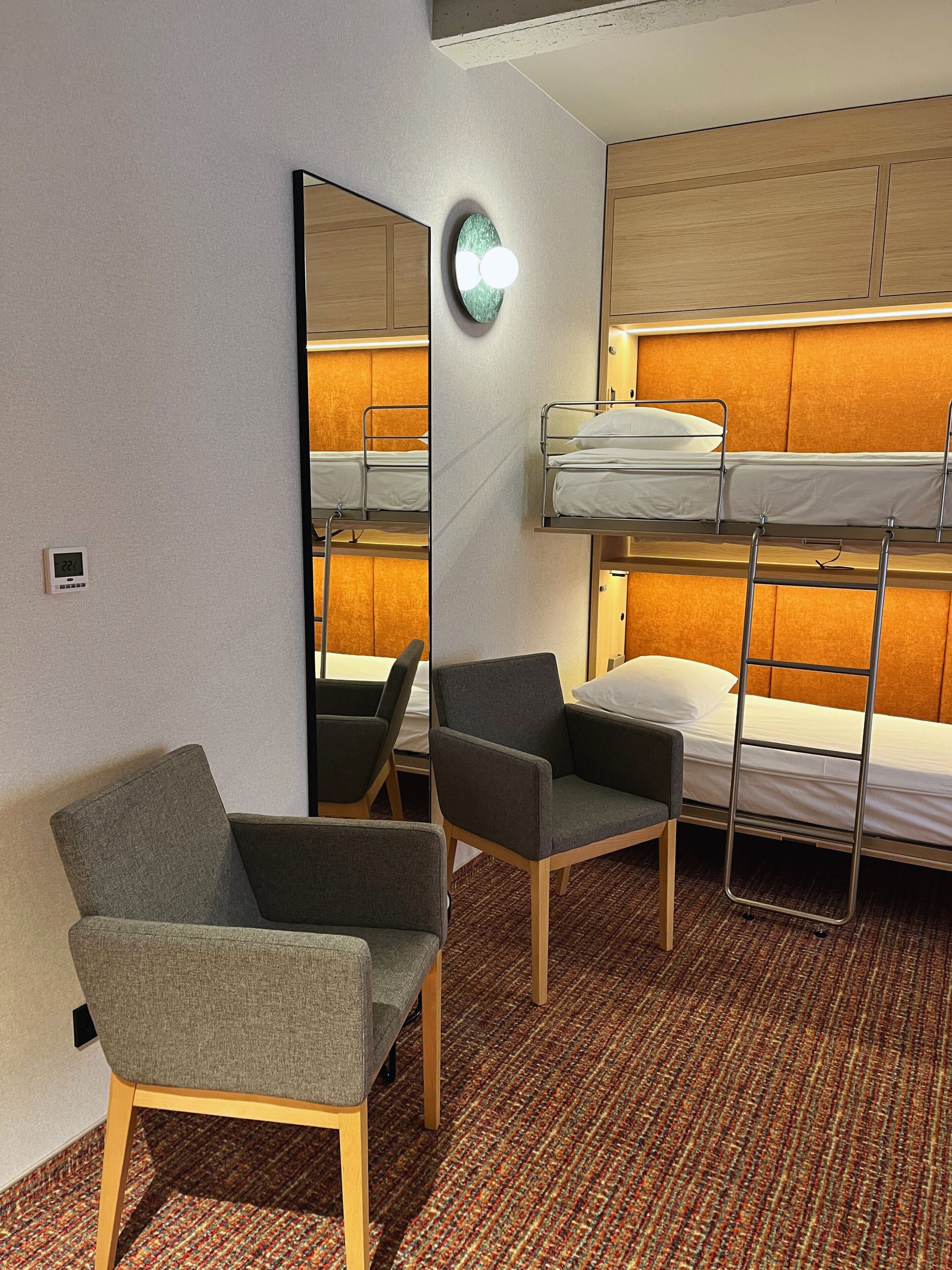 A hotel room with bunk beds, a mirror, two chairs, and a carpeted floor.