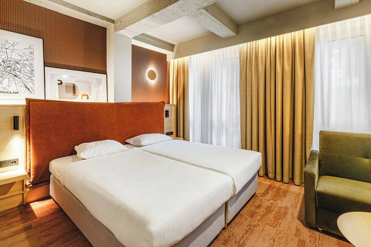 A hotel room with a large bed, a green armchair, and a desk with a lamp.