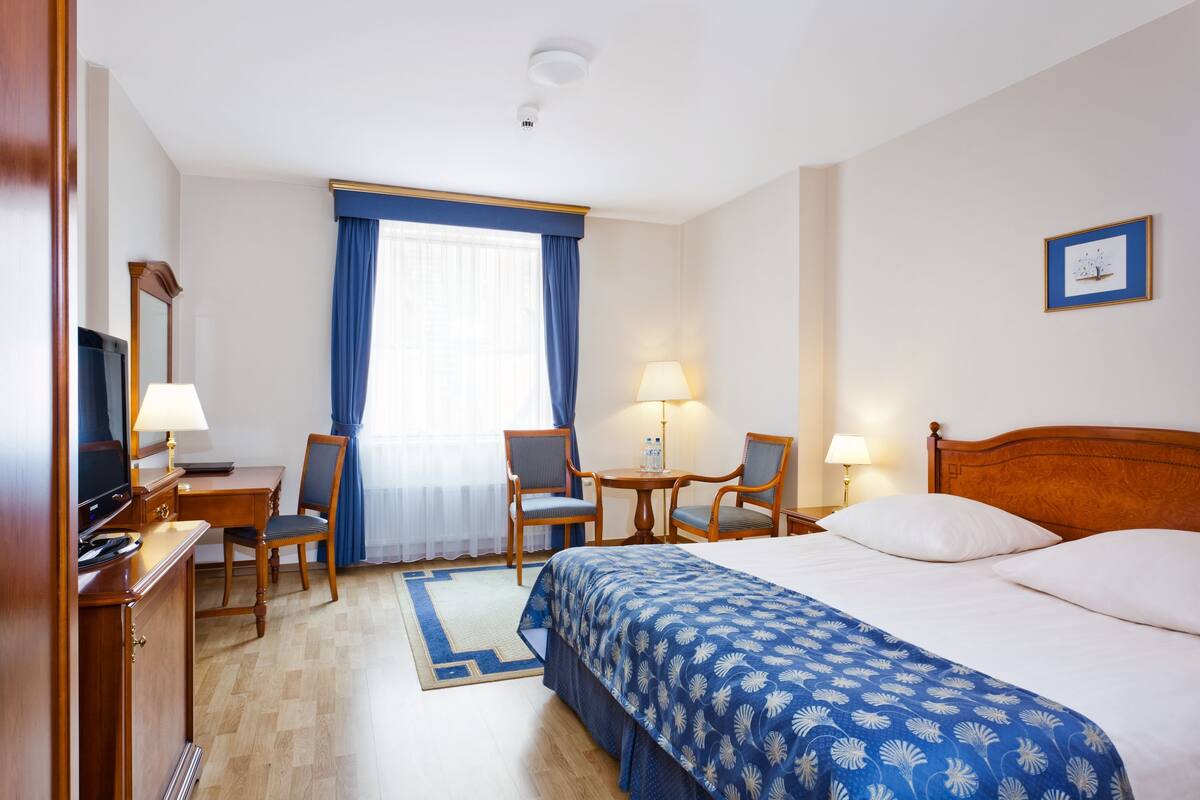 A hotel room with a bed, two chairs, a desk with a computer, a lamp, and a window with curtains.