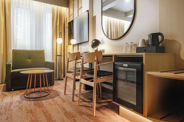 Une chambre d’hôtel équipée d’un canapé, de deux fauteuils, d’une petite table, d’une télévision, d’un miroir, d’une machine à café et d’un mini-réfrigérateur.