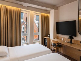 Habitación de hotel con dos camas, un escritorio, televisor y vistas a un edificio de ladrillo.