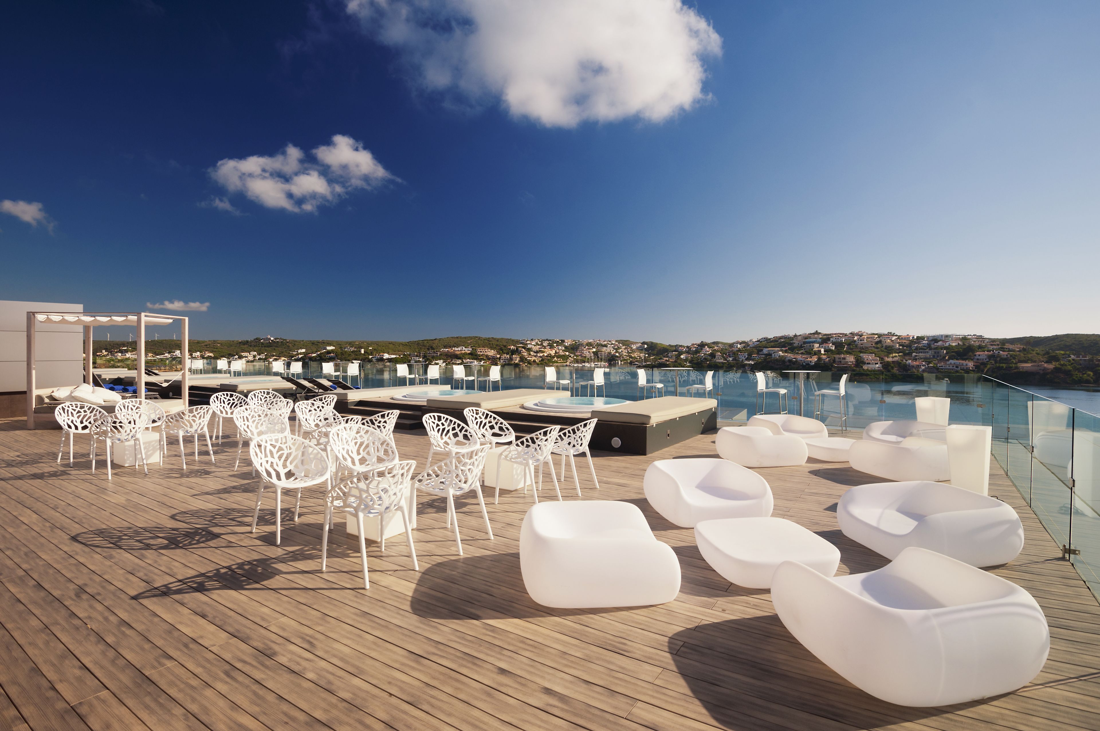 A rooftop terrace with white chairs and sofas, a pool, and a view of a town in the distance.