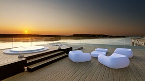 Une terrasse en bois avec des chaises longues blanches et un jacuzzi, offrant une vue sur une étendue d’eau au coucher du soleil.