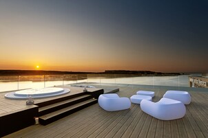 Una terraza de madera con sillones blancos y un jacuzzi, con vistas a un cuerpo de agua al atardecer.