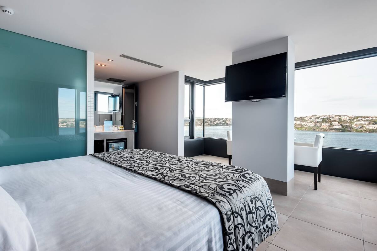 Une chambre d’hôtel moderne dotée d’un grand lit, d’une télévision, d’un balcon avec vue sur la mer et d’une salle de bain avec un grand miroir.