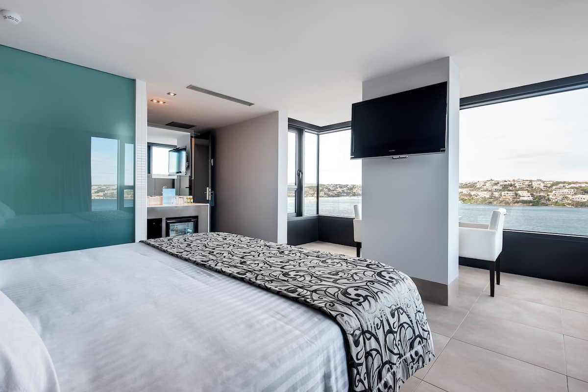 A modern hotel room with a large bed, a TV, a balcony with a view of the sea, and a bathroom with a large mirror.