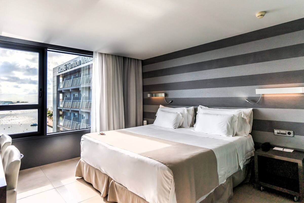 Une chambre d’hôtel avec un lit, un bureau et une fenêtre donnant sur un immeuble et une plage.