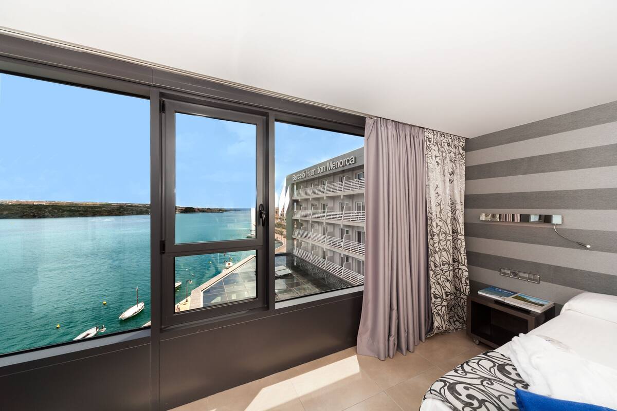 Une chambre d’hôtel dotée d’une grande fenêtre donnant sur un plan d’eau et sur un bâtiment appelé « Barceló Hamilton Menorca ».
