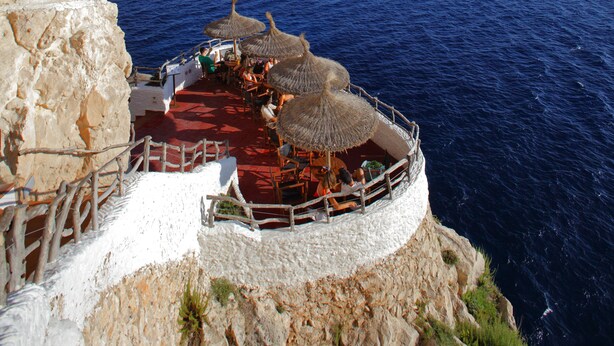 Une falaise en bord de mer, surmontée d’une construction aux toits rouges et agrémentée de parasols en chaume, d’où l’on a une vue sur l’océan.