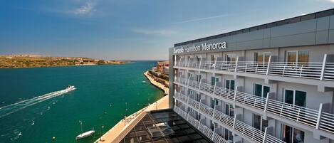 A modern hotel with a name sign, a balcony with railings, and a view of the sea and boats.