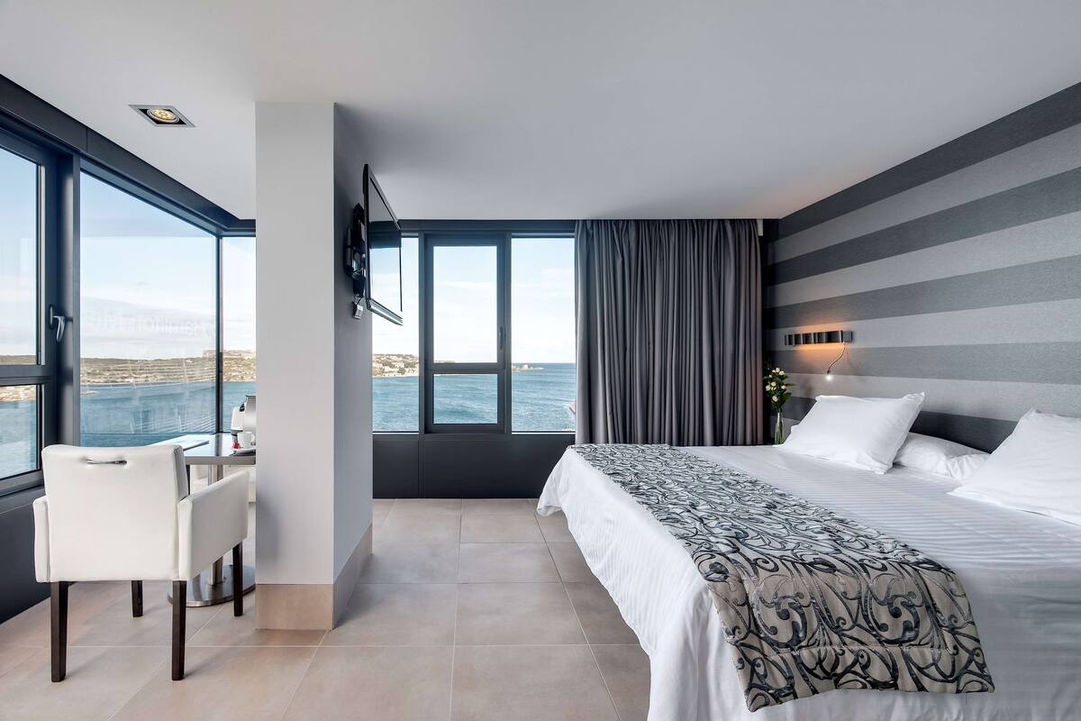 Une chambre d’hôtel moderne dotée d’une grande fenêtre, d’un lit avec une literie à motifs, d’un bureau avec une chaise et offrant une vue sur la mer.