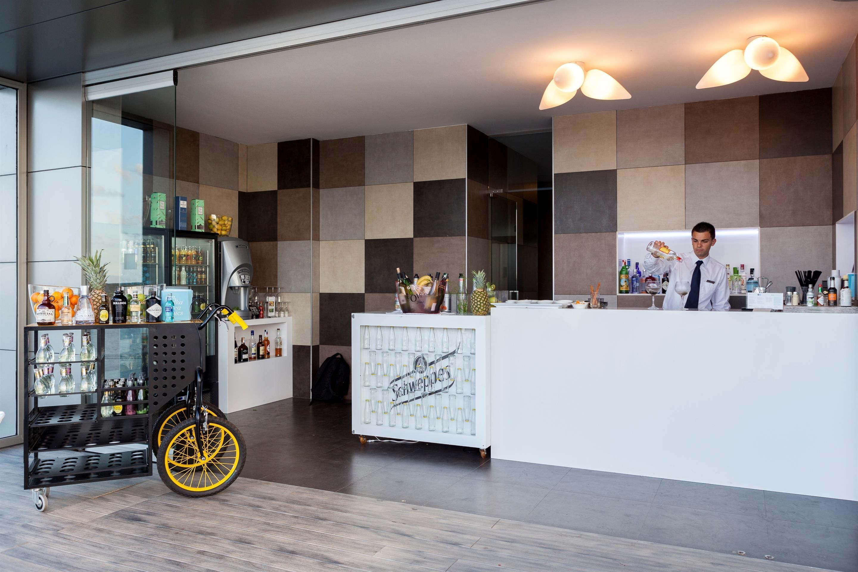 A modern bar area with a checkered wall design, a wheelchair-accessible cart with beverages, and a bartender preparing drinks.