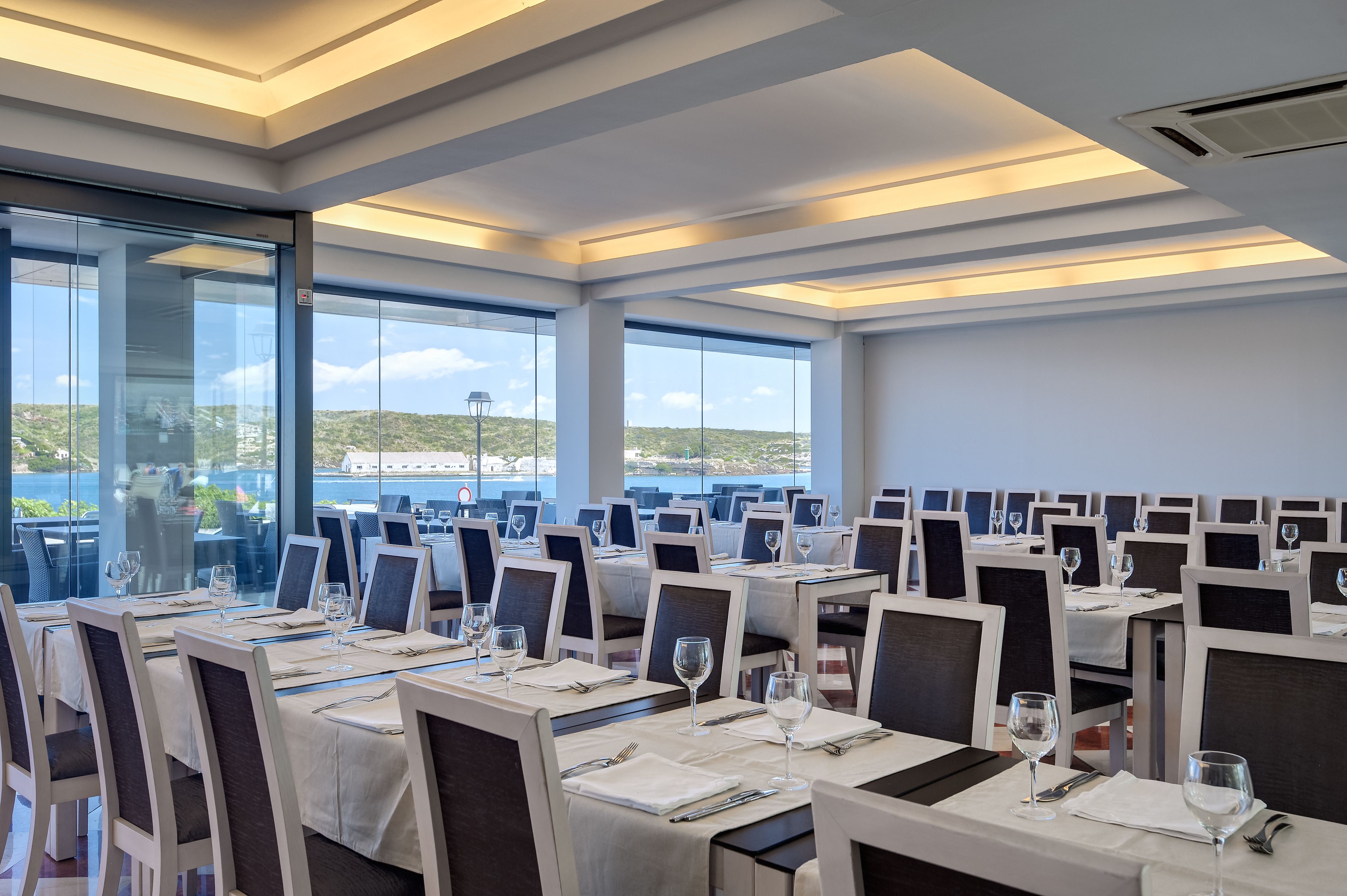 Un restaurante con mesas dispuestas para comer, ventanas amplias y vista al mar.