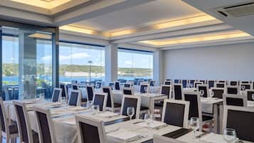 Un restaurante con mesas dispuestas para comer, ventanas amplias y vista al mar.