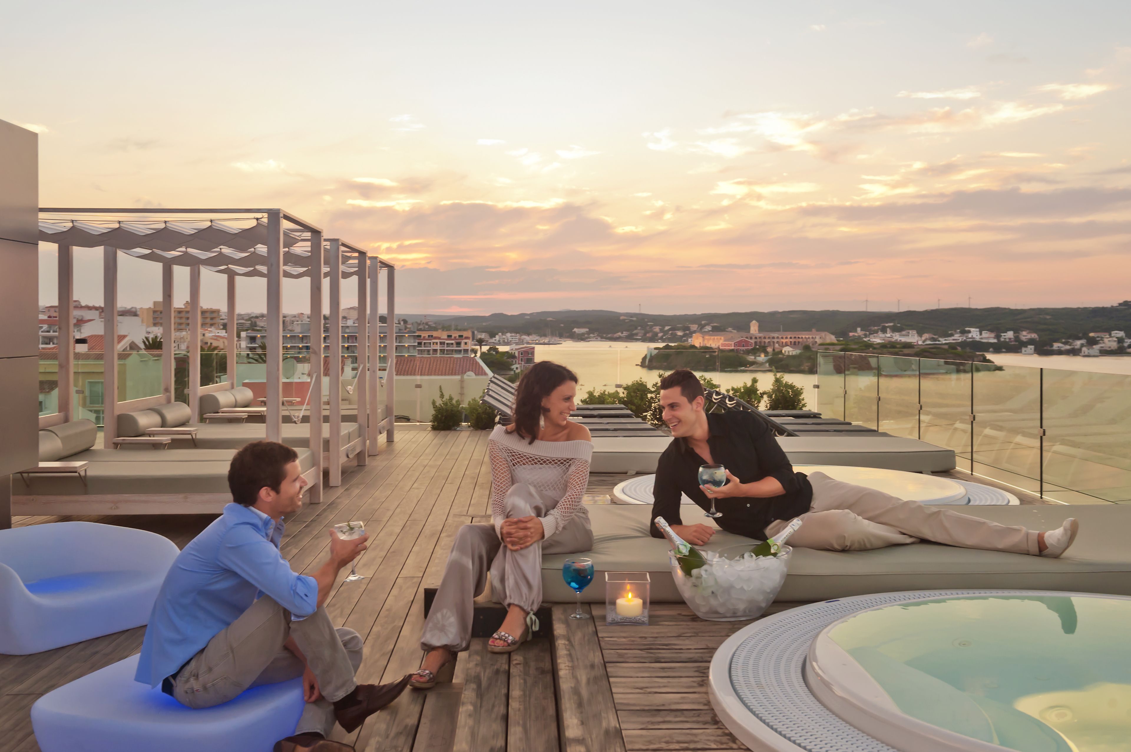 A rooftop terrace with seating areas, a hot tub, and a view of a cityscape at sunset.
