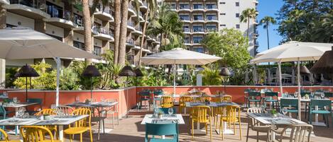An outdoor dining area with tables and chairs, surrounded by palm trees and a building with balconies.