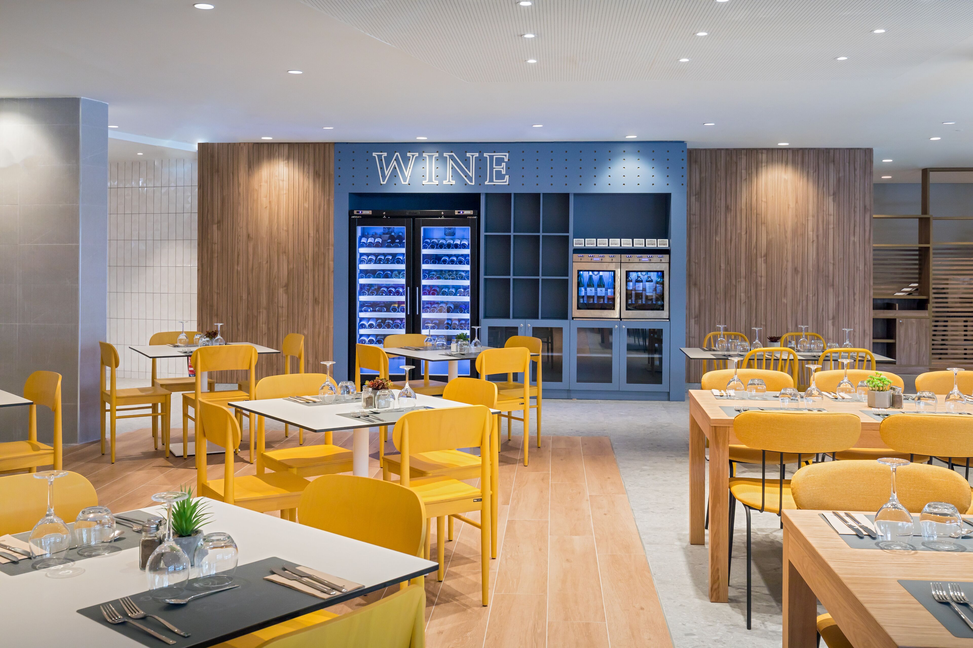 A modern restaurant with yellow chairs and tables, a wine display, and a blue wall with the word "WINE".