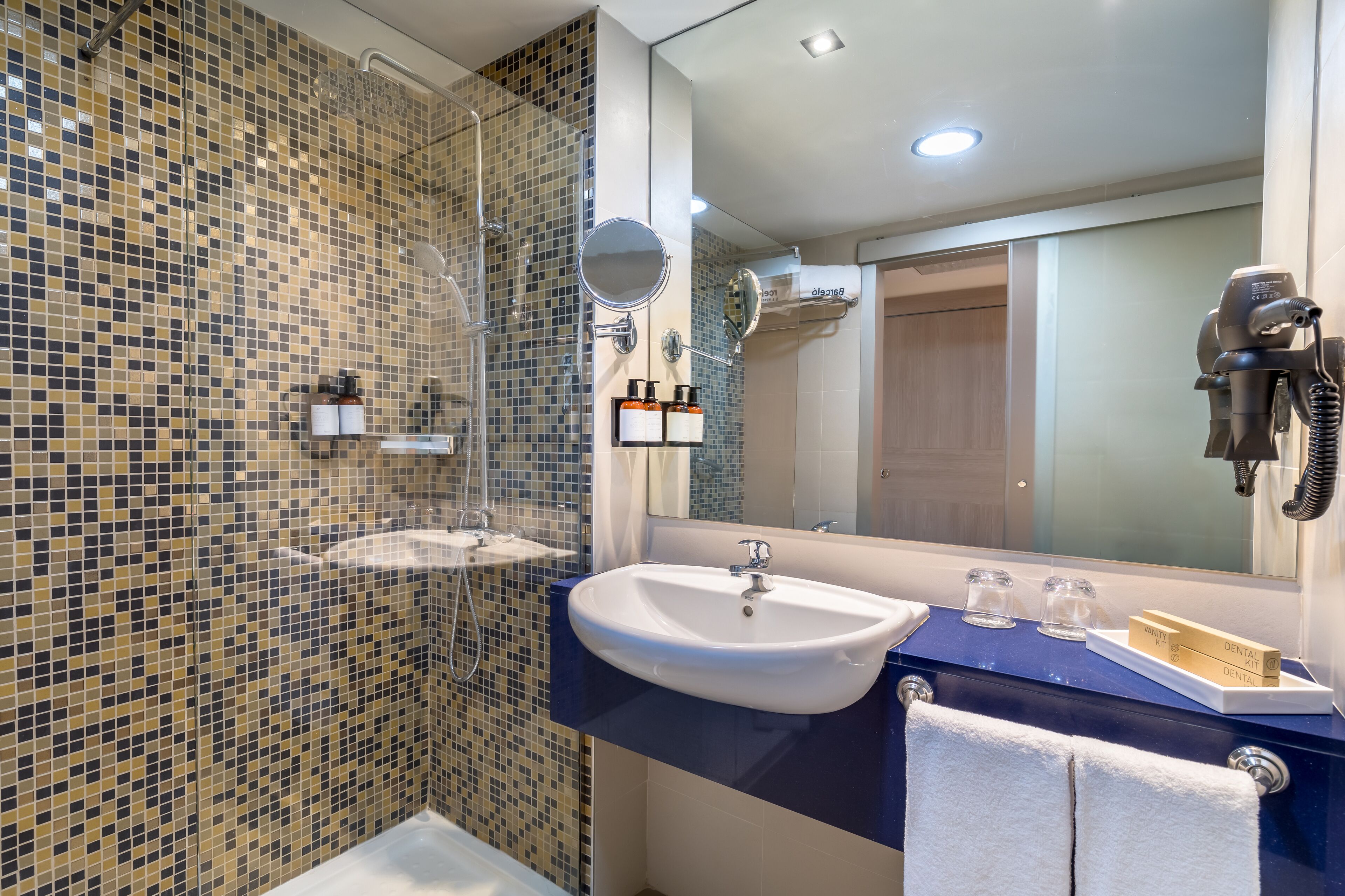 Un baño con pared de ducha de mosaico, un lavamanos con encimera azul, un espejo, un secador de pelo y un perchero para toallas.
