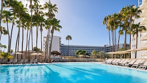 Una piscina con sillas de descanso y sombrillas, rodeada de palmeras y un edificio de hotel al fondo.