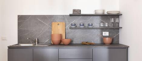 Una cocina moderna con armarios grises, salpicadero de mármol, tabla de cortar de madera y una repisa con cuencos y vasos.