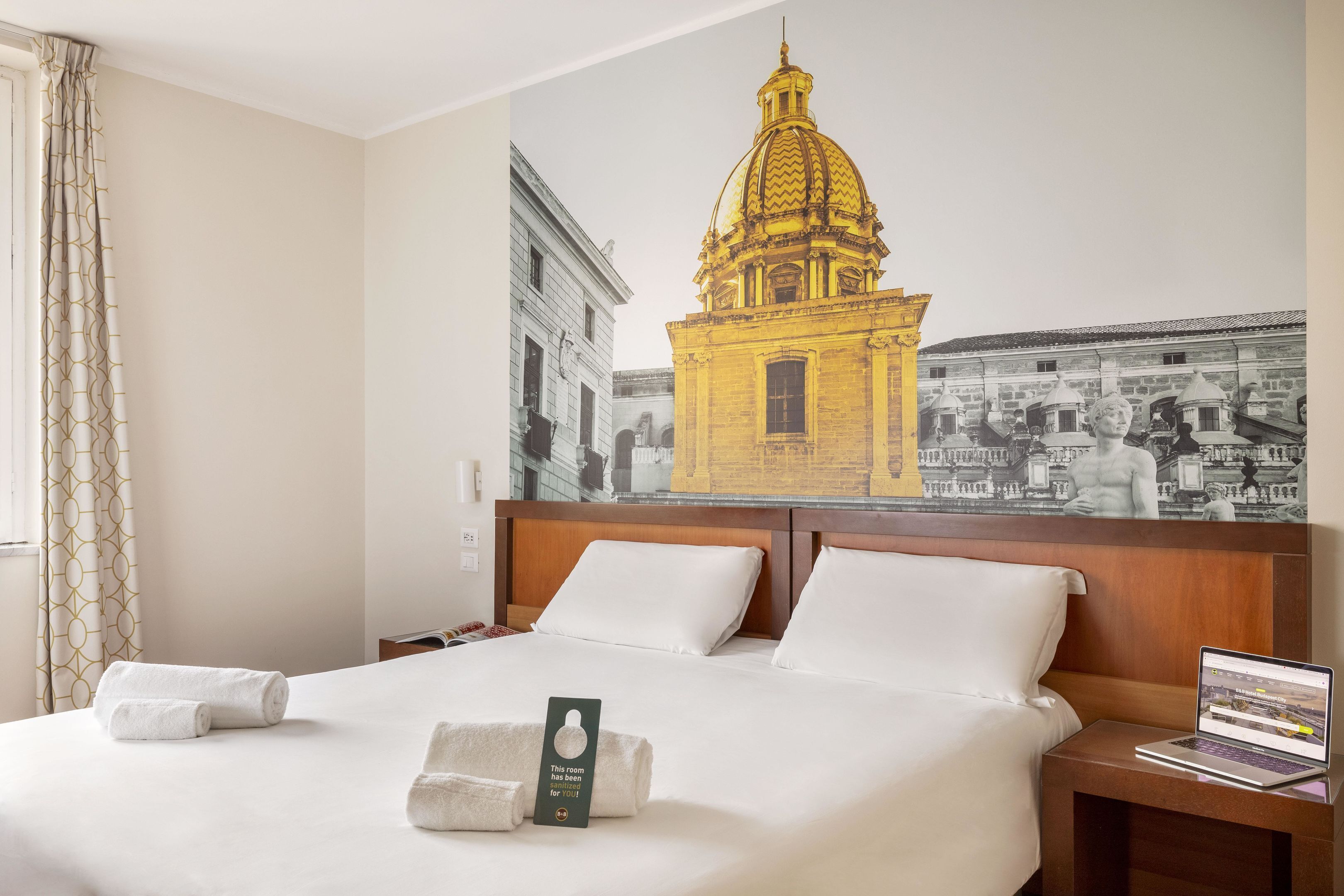Ein Hotelzimmer mit einem großen Bett, einem Laptop auf einem Nachttisch und einem Wandbild eines Gebäudes.
