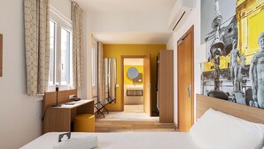 Habitación de hotel con cama, escritorio, silla, mesita pequeña y baño visible a través de una puerta abierta.