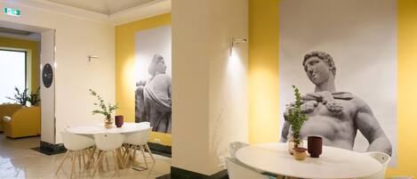 A hotel lobby with a round table and chairs, a large mural of a classical statue, and a yellow accent wall.
