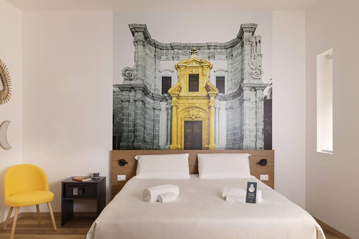 A hotel room with a large bed, a yellow chair, a small bedside table, and a wall mural of a building.