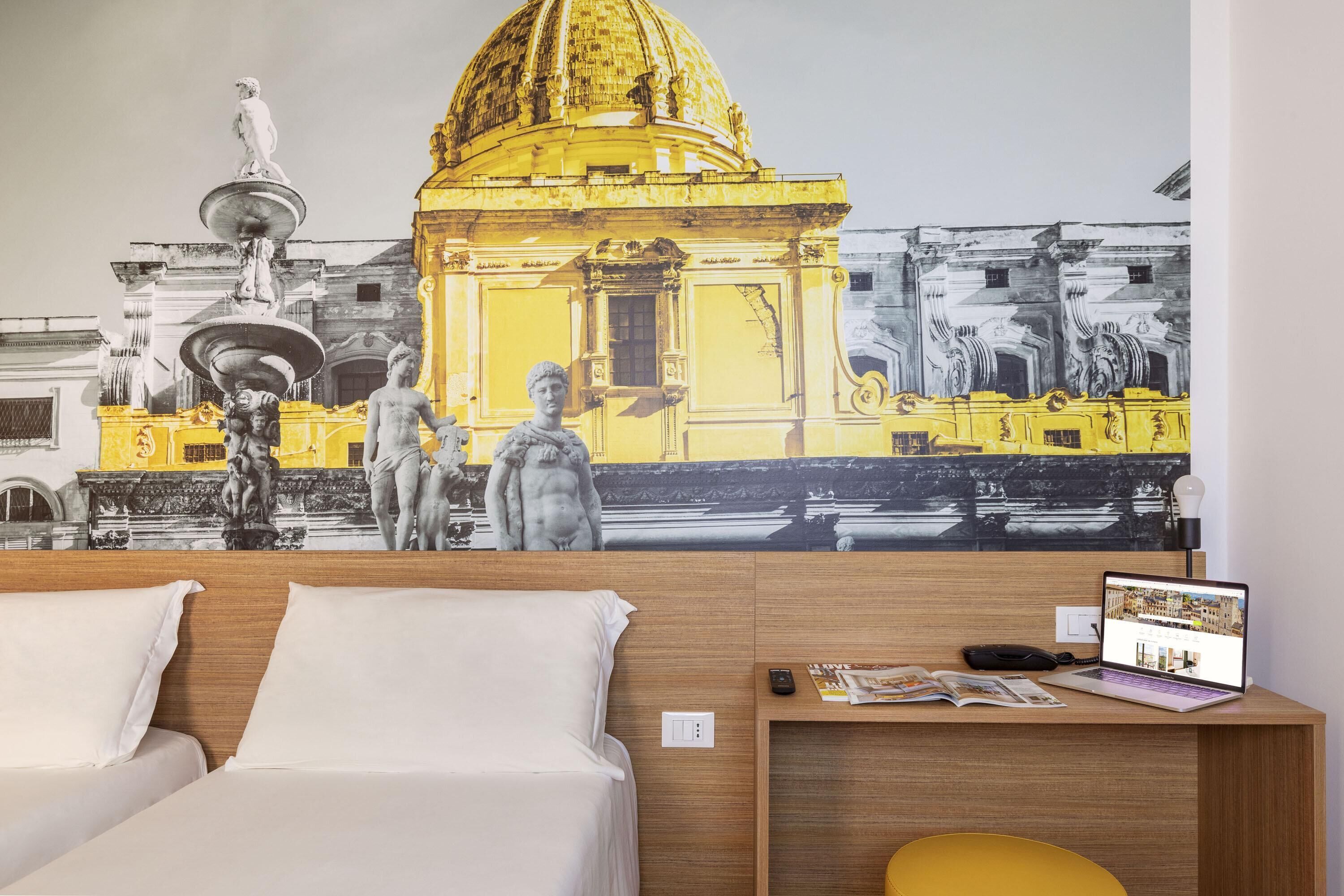 Habitación de hotel con cama, escritorio y una computadora portátil, junto a un mural de un edificio histórico.