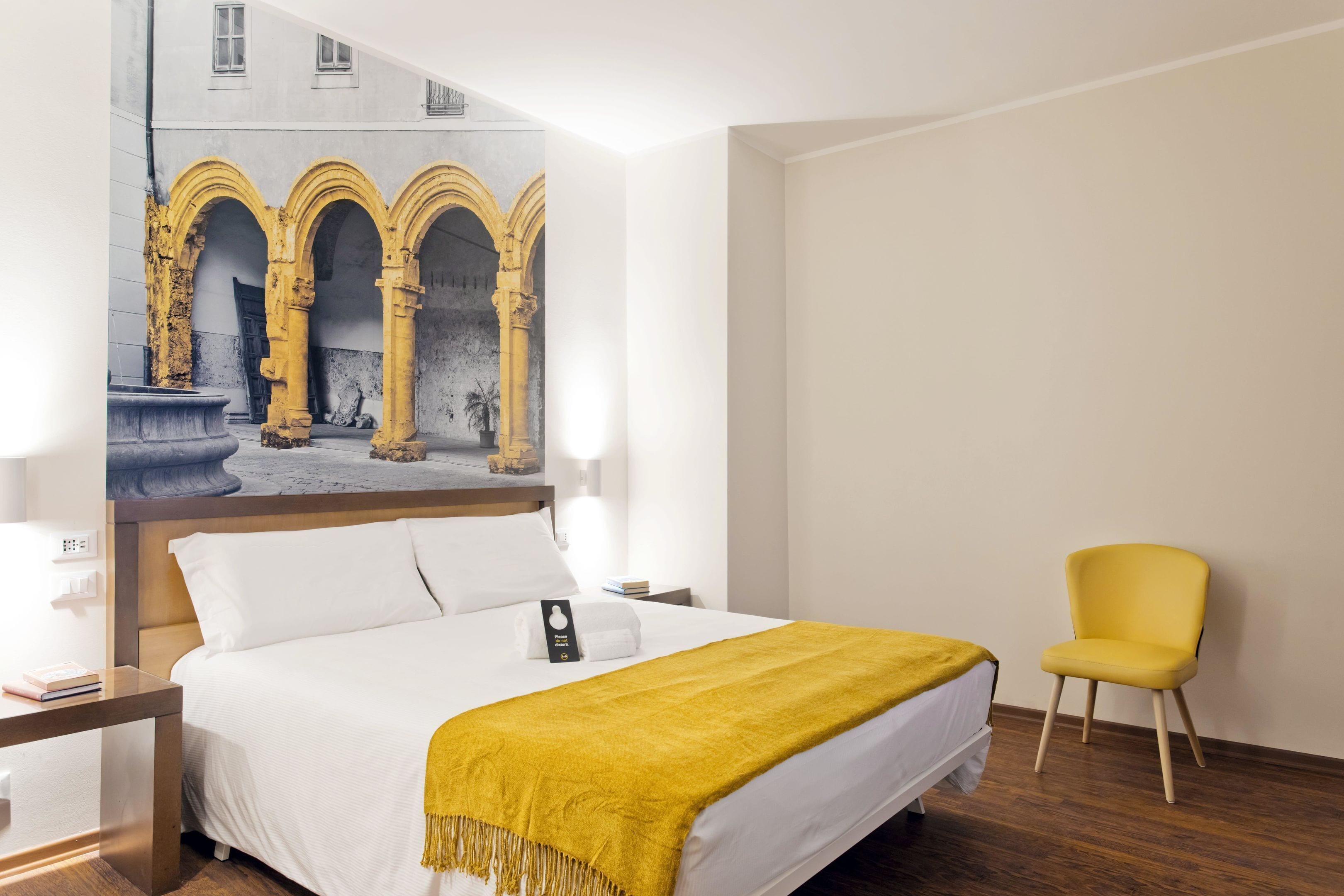 A hotel room with a bed, a yellow chair, and a painting of arches on the wall.