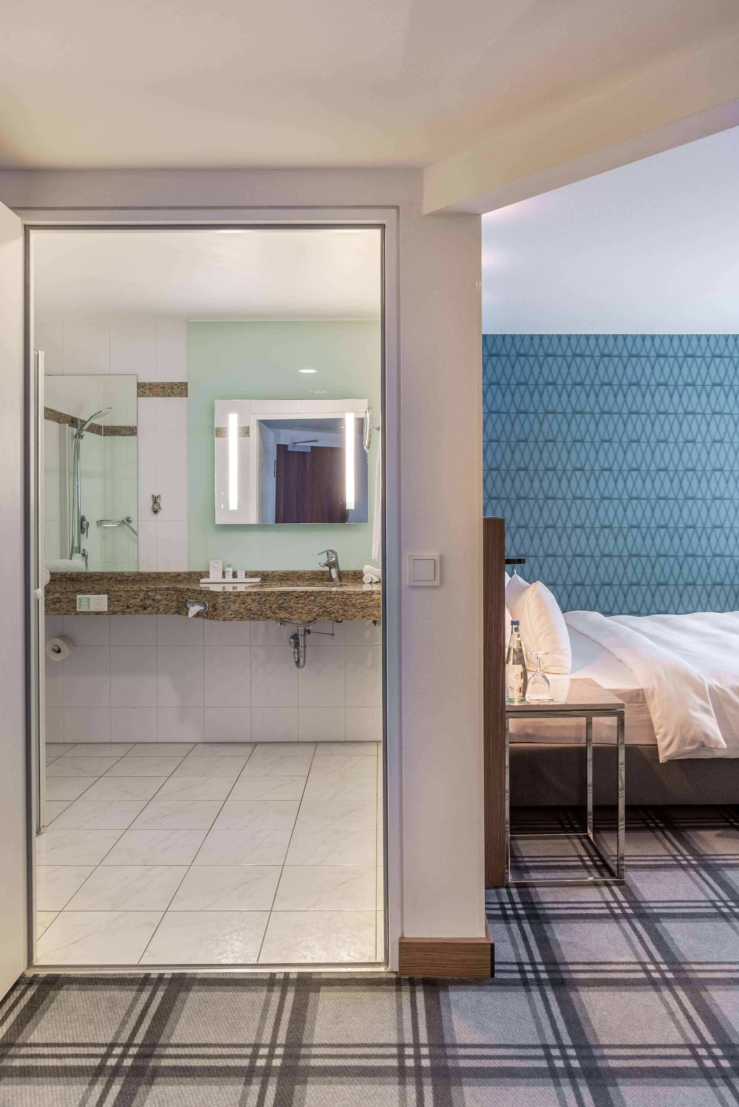 A hotel room with a bathroom featuring a glass door, a sink with a granite countertop, and a mirror. The room has a bed with white bedding and a blue patterned wall.