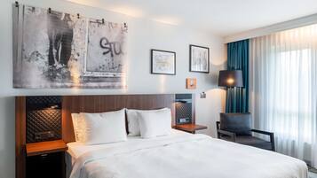 Une chambre d’hôtel avec un grand lit, une tête de lit à motifs matelassés, des tables de chevet, une lampe et des œuvres d’art accrochées au mur.