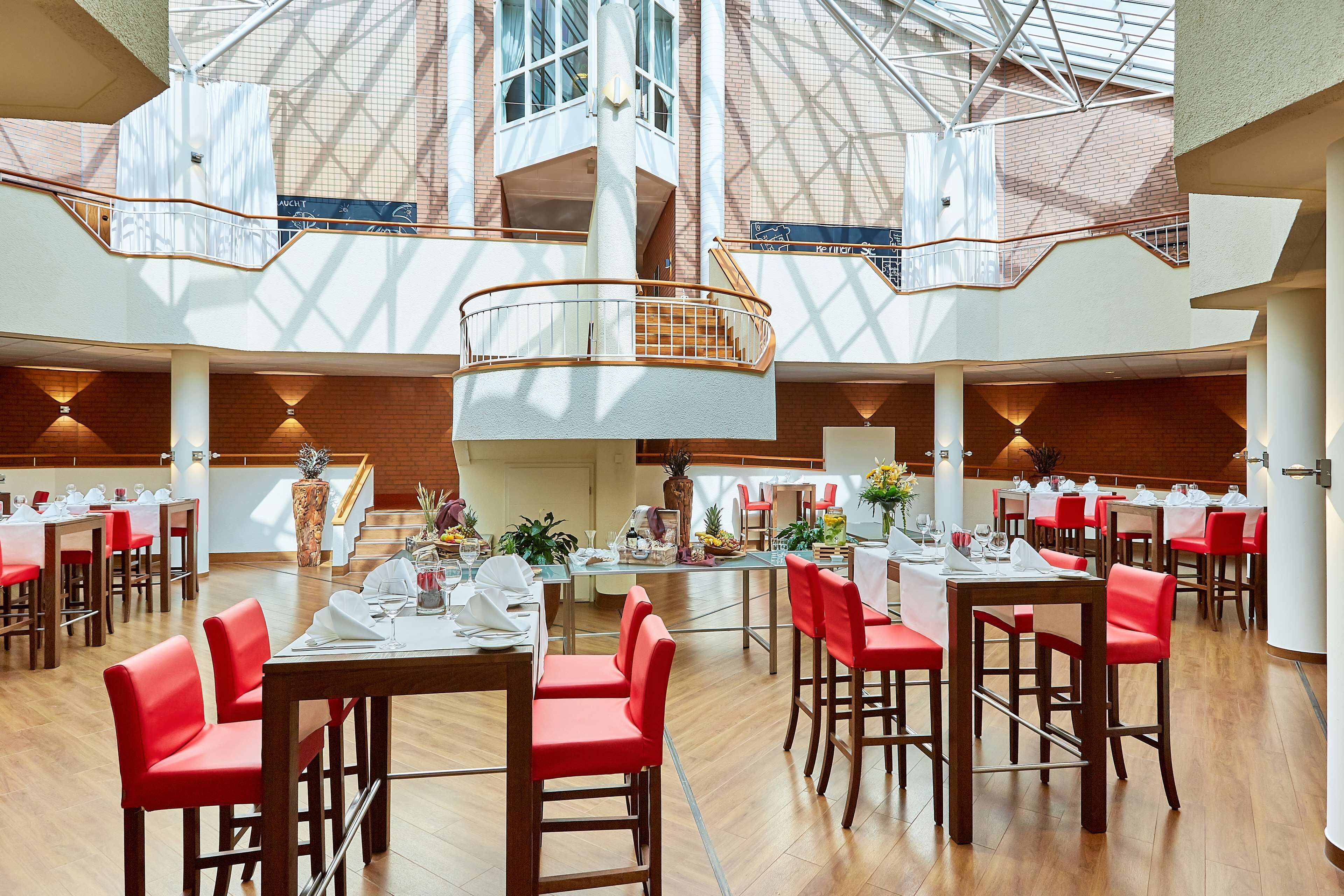 Un vaste espace dédié à la salle à manger, avec de nombreuses tables et des chaises rouges, un escalier central et un plafond haut doté de lucarnes.