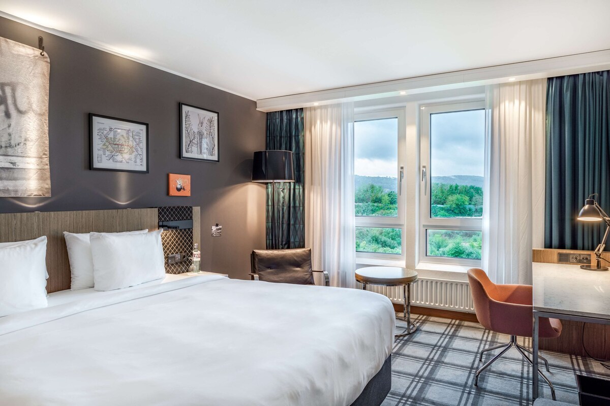 Quarto de hotel com uma cama grande, uma escrivaninha, uma cadeira, uma mesinha e vista para o verde pela janela.