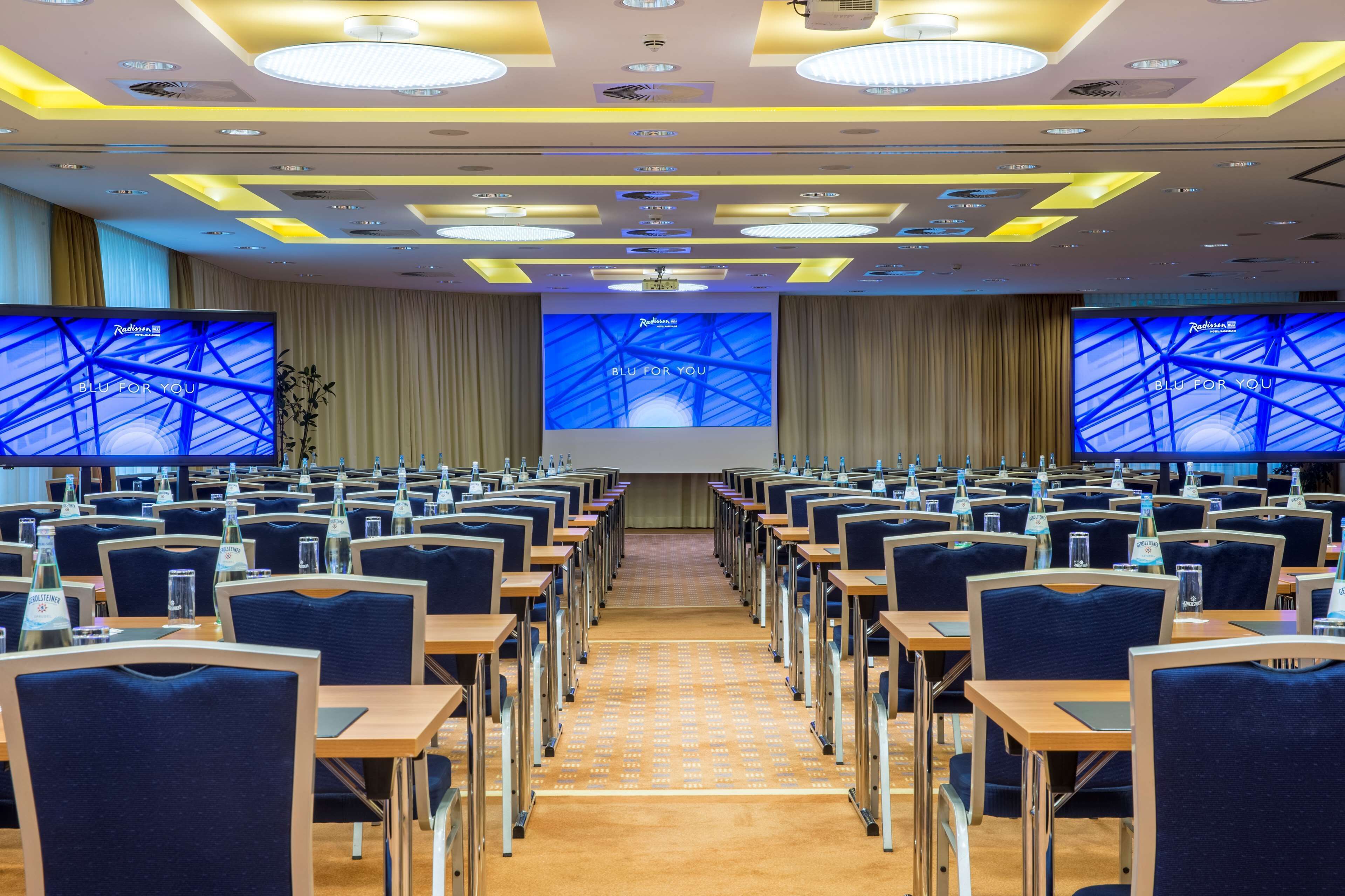 Ein Konferenzraum mit Tischen und Stühlen, großen Bildschirmen an den Wänden und einem modernen Deckengestaltungsstil.