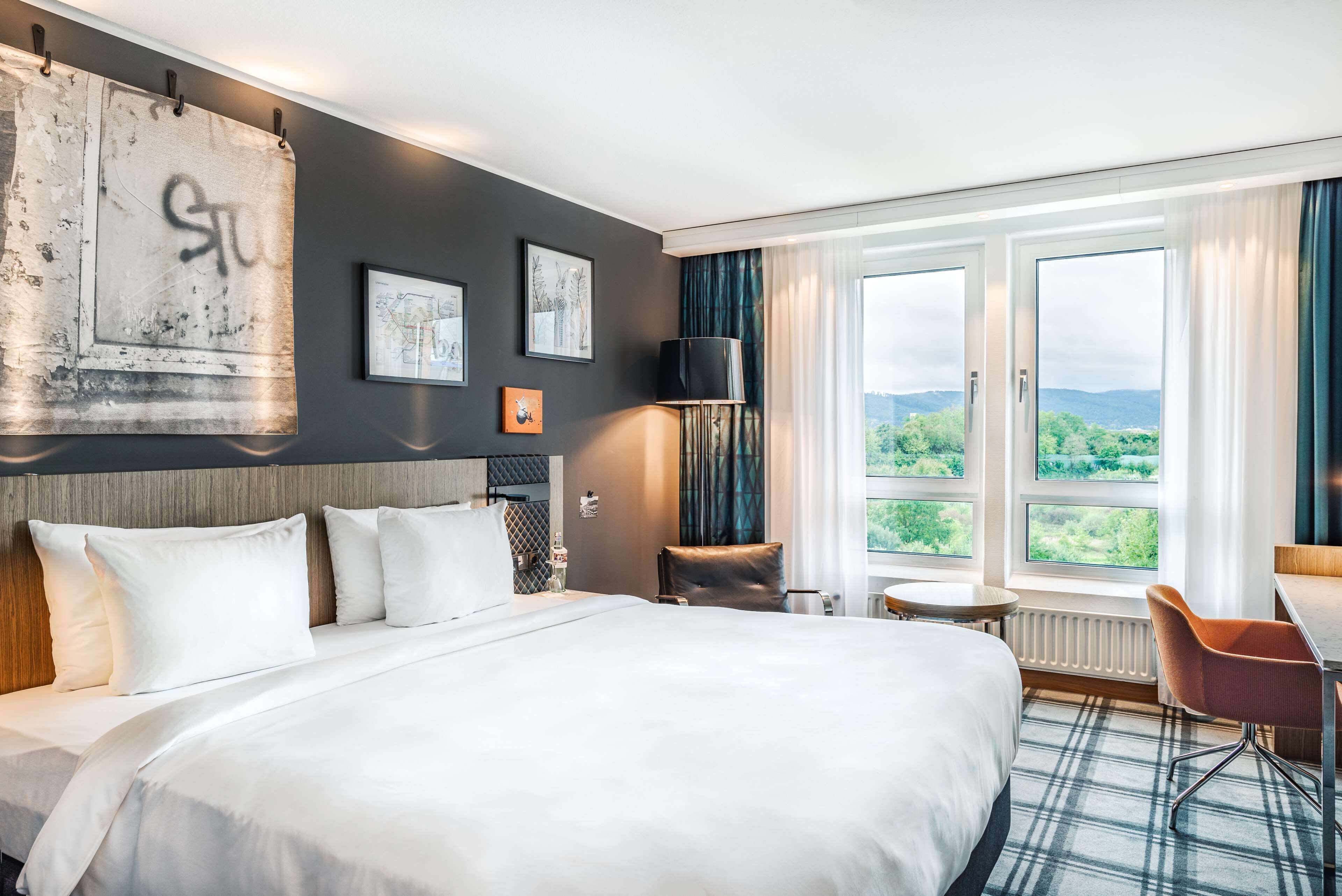 A hotel room with a large bed, a view of greenery through the windows, a desk, and a chair.