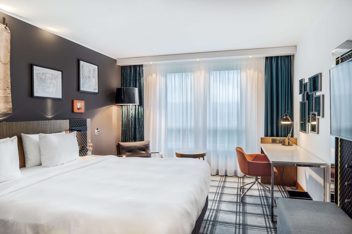 A hotel room with a large bed, a desk, a chair, and a view of the city through the window.