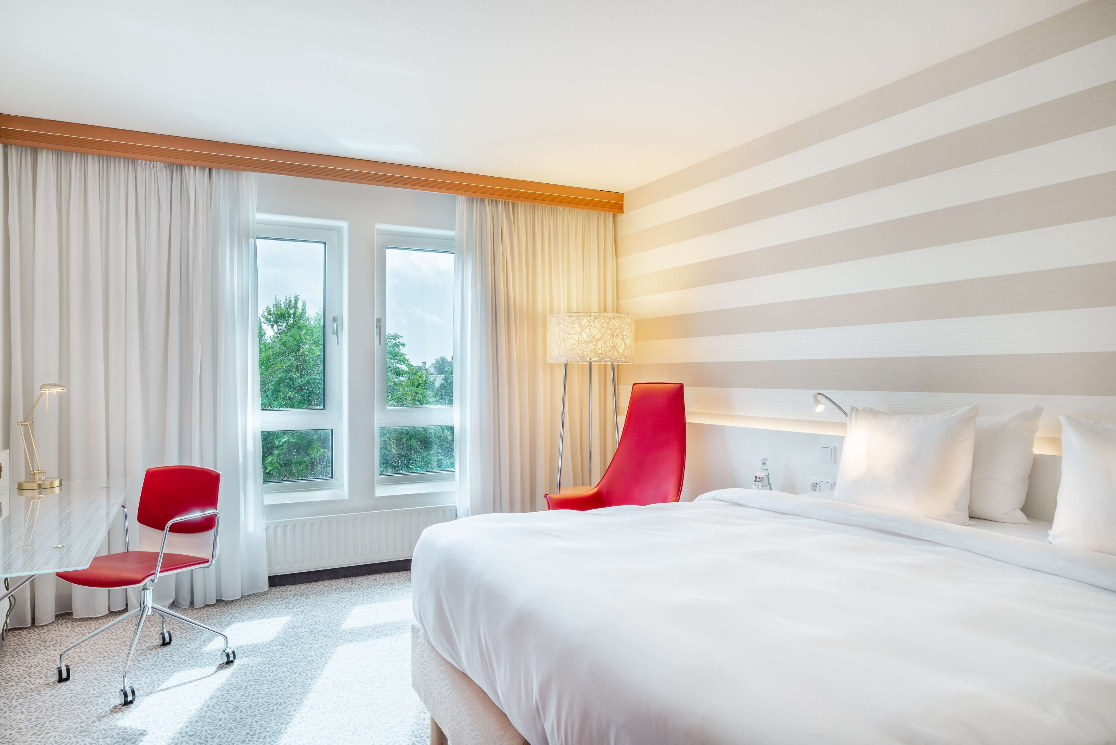 A hotel room with a large bed, a desk with a chair, a lamp, and a view of greenery outside.